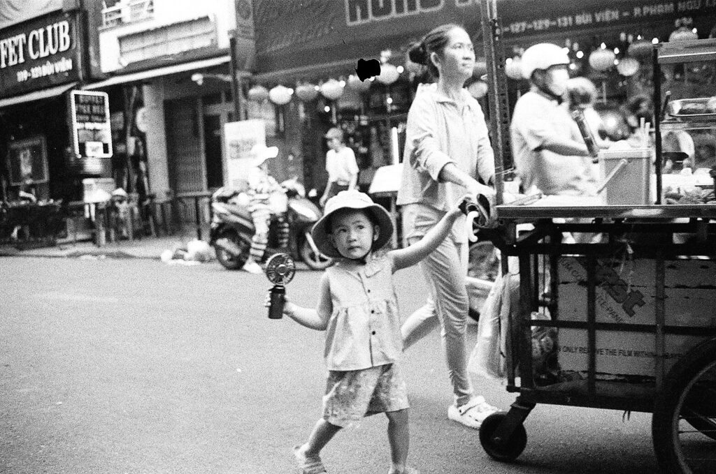 About A small child walking along a busy street with a woman behind, shot on black and white film