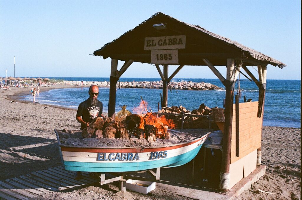 About Fish grilling over an open fire beside a painted wooden boat, with the sea behind