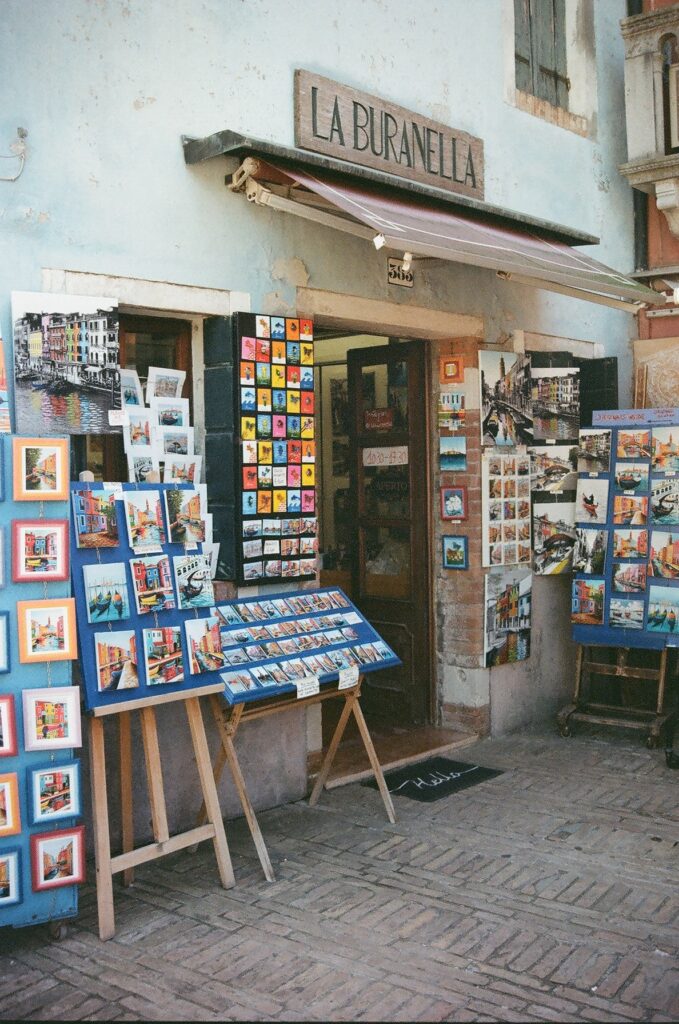 Home A small art shop with colourful paintings on easels outside its door in Burano, Venice, shot on film