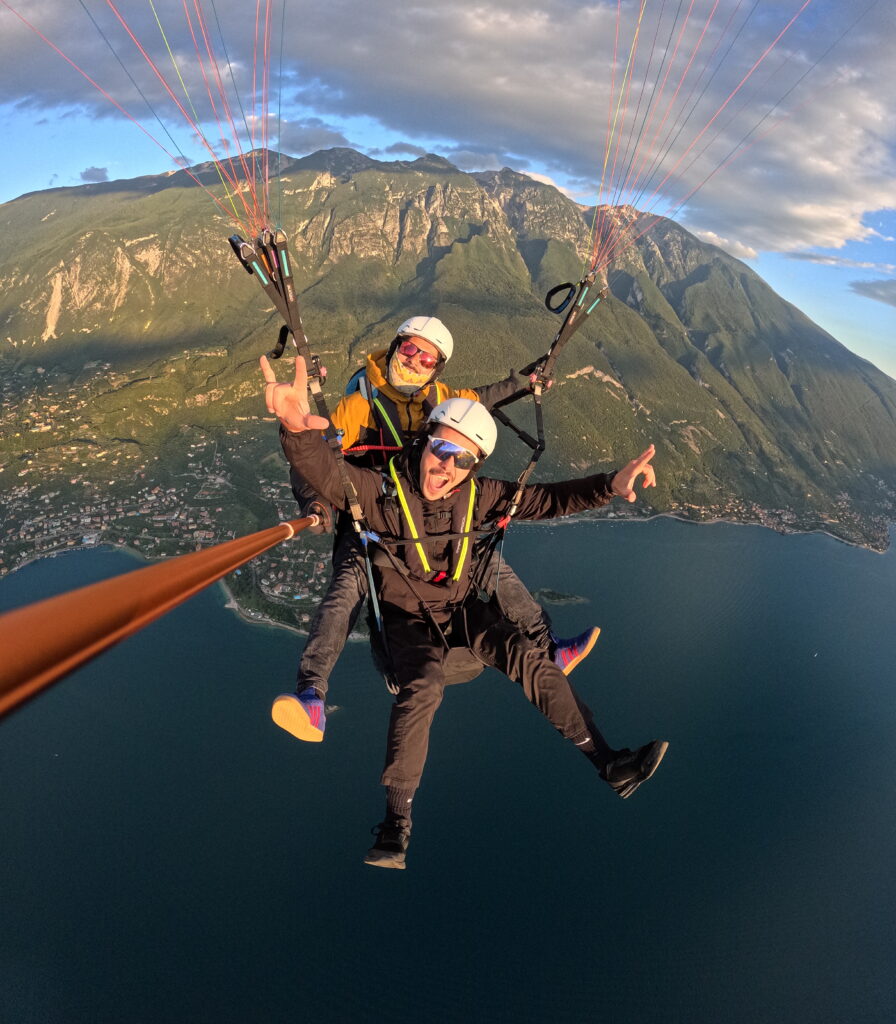 Home Two people flying a tandem paraglider high above a lake, with mountains behind them